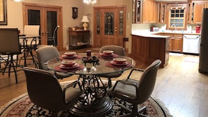 A dining area with a round glass table surrounded by chairs, a chandelier, wooden cabinets, and a kitchen area with wooden cabinets and a refrigerator.
