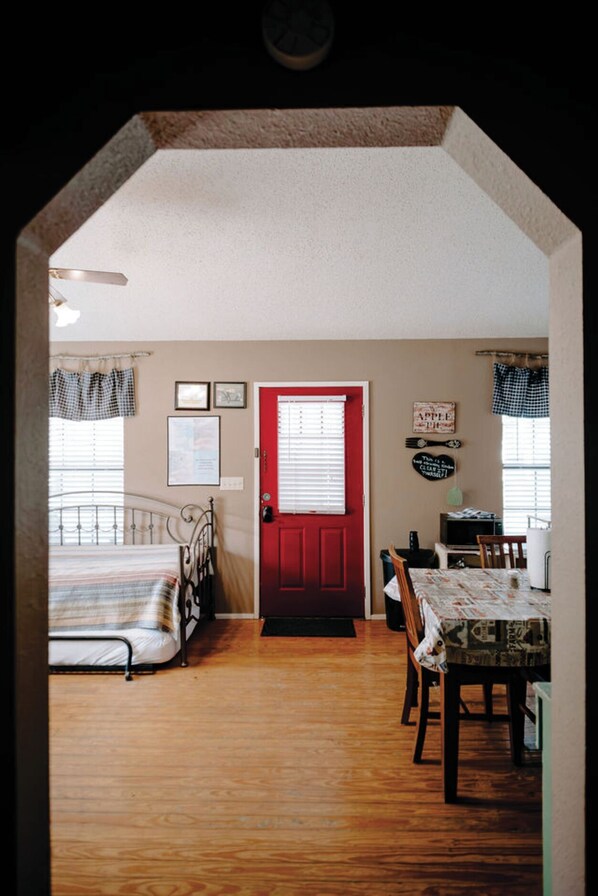 A room with a bed, a dining table with chairs, a red door, and a window with blinds.