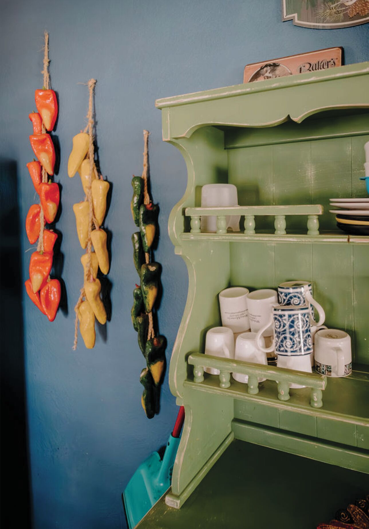 Una cocina con un armario verde que contiene tazas blancas y azules, y una pared con pimientos colgando.
