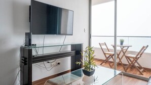 Ein modernes Wohnzimmer mit einem flachbildfernseher an einem Glastisch, einem kleinen Tisch mit Stühlen und einer Topfpflanze.