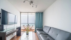 Ein modernes Wohnzimmer mit grauem Sofa, einem Flachbildfernseher, einem Glastisch und Meerblick durch große Fenster.