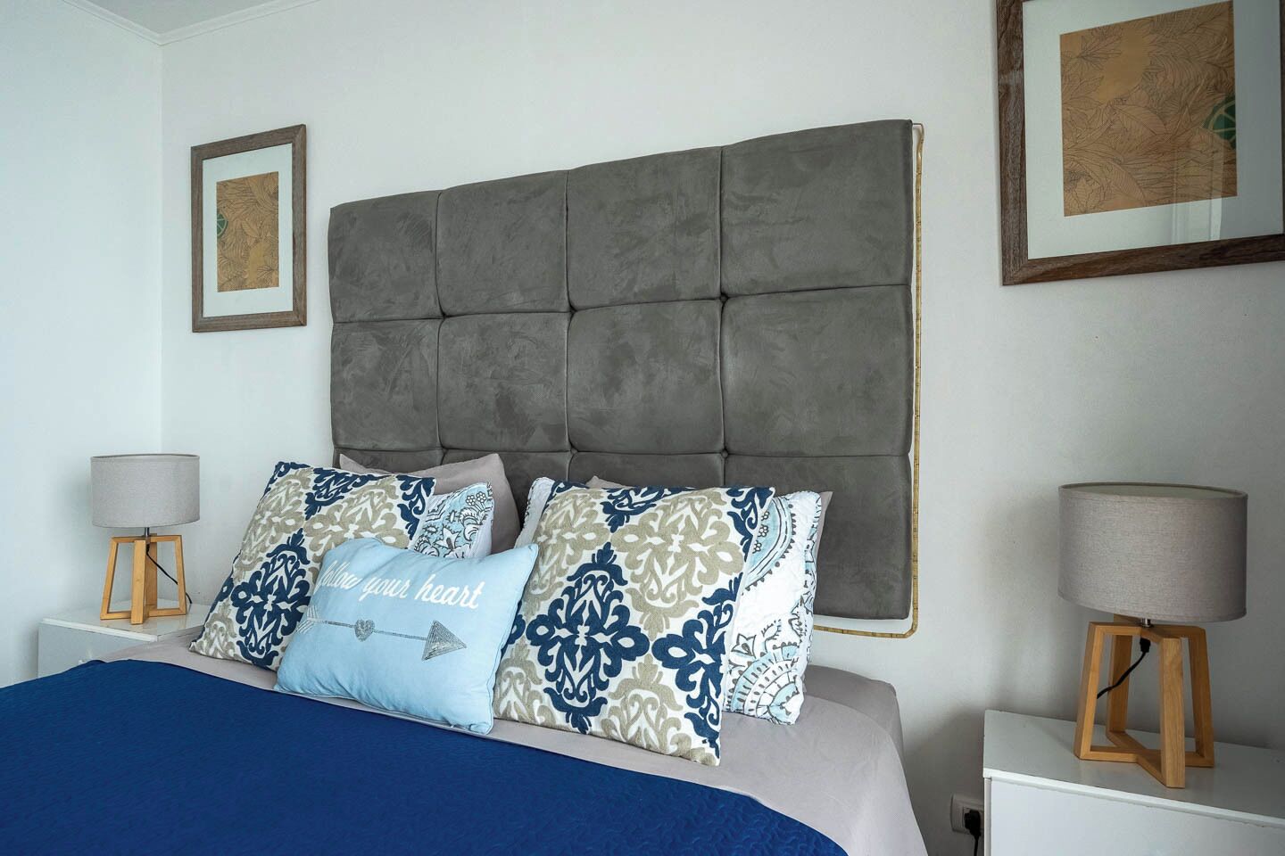 Une chambre à coucher avec une tête de lit capitonnée, des coussins à motifs, une couvre-lit bleue et des lampes de chevet posées sur des supports en bois.