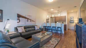 A modern living room with a sofa, coffee table, and a rug. There is a kitchen area with bar stools and a dining table.