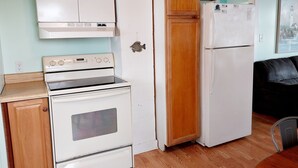 Una cucina compatta con piano cottura, forno e frigorifero bianchi, pensili in legno e una piccola decorazione a forma di pesce sul muro.