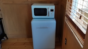 A compact kitchenette with a microwave and refrigerator, wooden walls, and a window with blinds.
