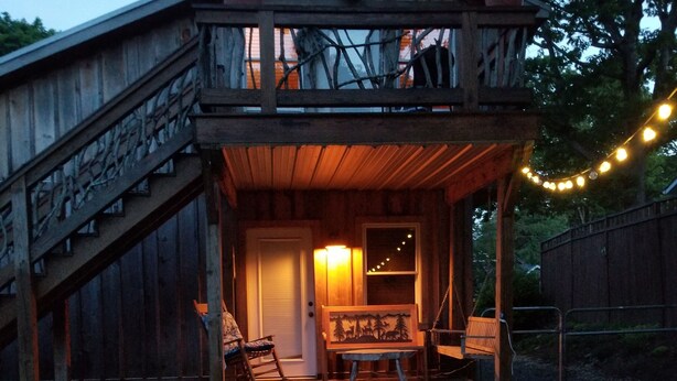 A wooden cabin with a covered porch, a staircase leading to an upper level, and a bench with a painting on the wall.