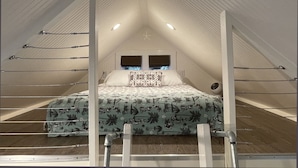 Un dormitorio en ático pequeño y triangular con cama, dos ventanas y un ventilador.