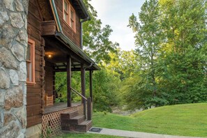 A wooden cabin with a stone facade, a covered porch, and a view of a lush green landscape. - Rippling Waters Retreat | Riverfront Rustic Cabin (Townsend)