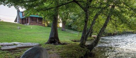 A riverside view with a stone bridge, a grassy area, and a cabin with a blue roof.