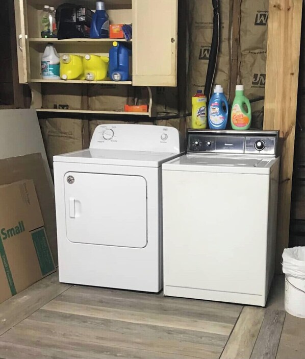 Un cuarto de lavandería con lavadora y secadora de carga frontal, un gabinete arriba con artículos de limpieza, y una caja de cartón con la etiqueta “Pequeña” en el suelo.