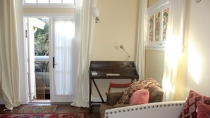 Ett mysigt rum med ett piano, en soffa med mönstrade kuddar och en utsikt genom en dörr ut mot ett utomhusområde.