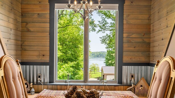 A room with a large window, wooden walls, a chandelier, a wooden bed, and a view of trees and a house.