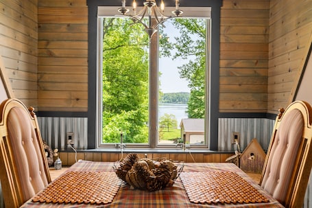 A room with a large window, wooden walls, a chandelier, a wooden bed, and a view of trees and a house.
