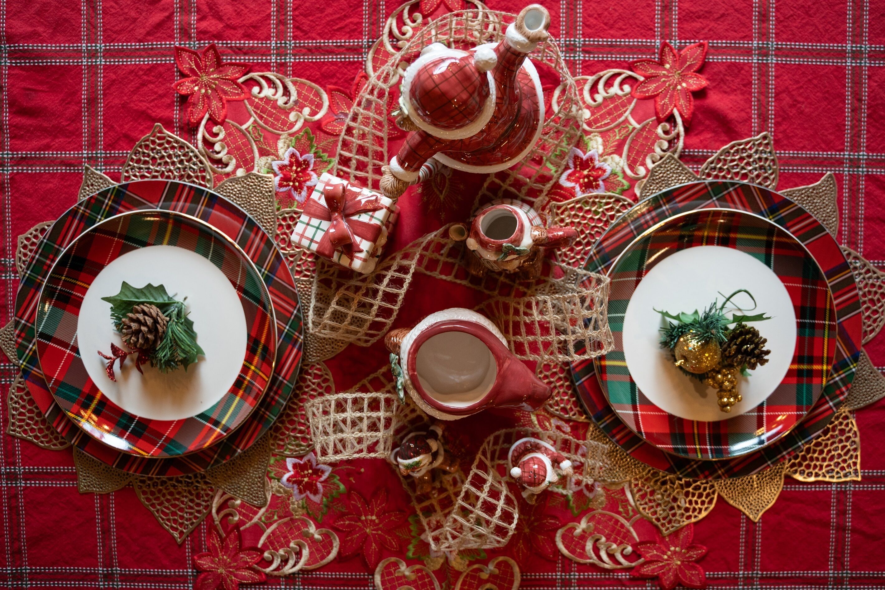 A table set with plaid-patterned plates, a teapot, and a cup, surrounded by festive decorations on a red tablecloth.