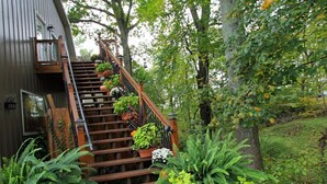 A wooden staircase leading to a house with a metal exterior, surrounded by lush greenery and potted plants.