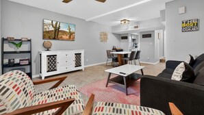 Un salon moderne avec un ventilateur de plafond, un coin repas avec une table et des chaises, une cuisine en arrière-plan et un mur gris orné d’un tableau représentant un cactus et d’une citation inspirante.