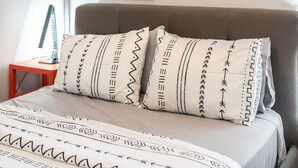 Un lit bien fait, avec une literie à motifs et des oreillers, une petite table de chevet en bois avec une lampe, et une tête de lit rembourrée de couleur grise.