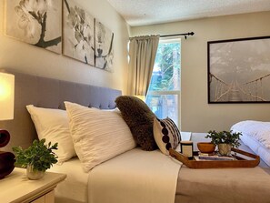A bedroom with a bed, pillows, a bedside table with a lamp, a window with curtains, and wall art.