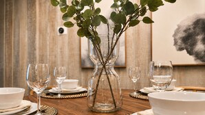 A dining table set with white plates, bowls, and wine glasses, with a clear glass vase containing green foliage in the center.
