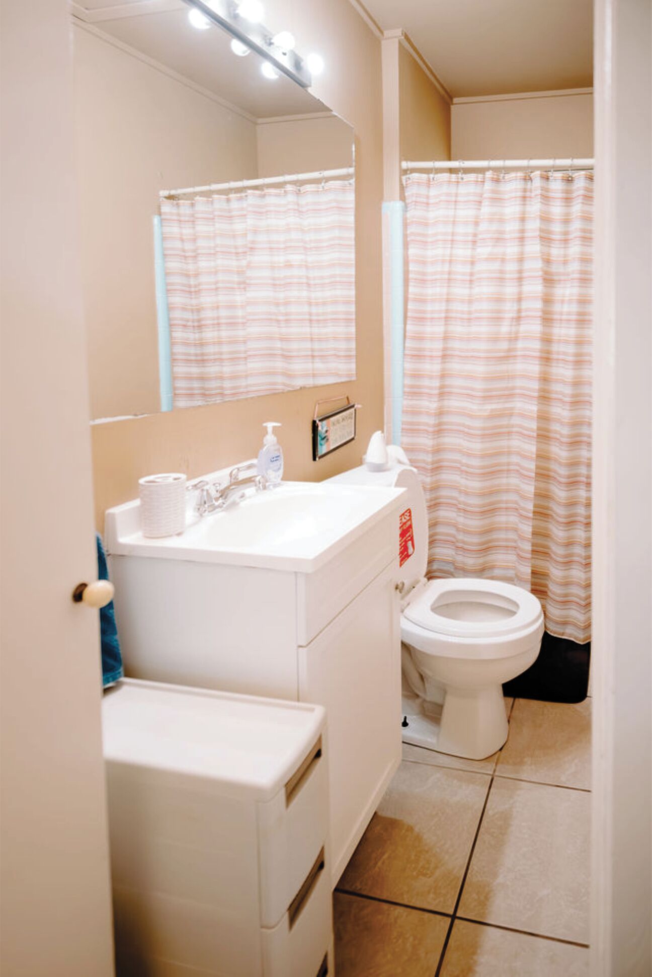 A bathroom with a white sink, mirror, and toilet. There is a striped shower curtain and a small shelf with toiletries.