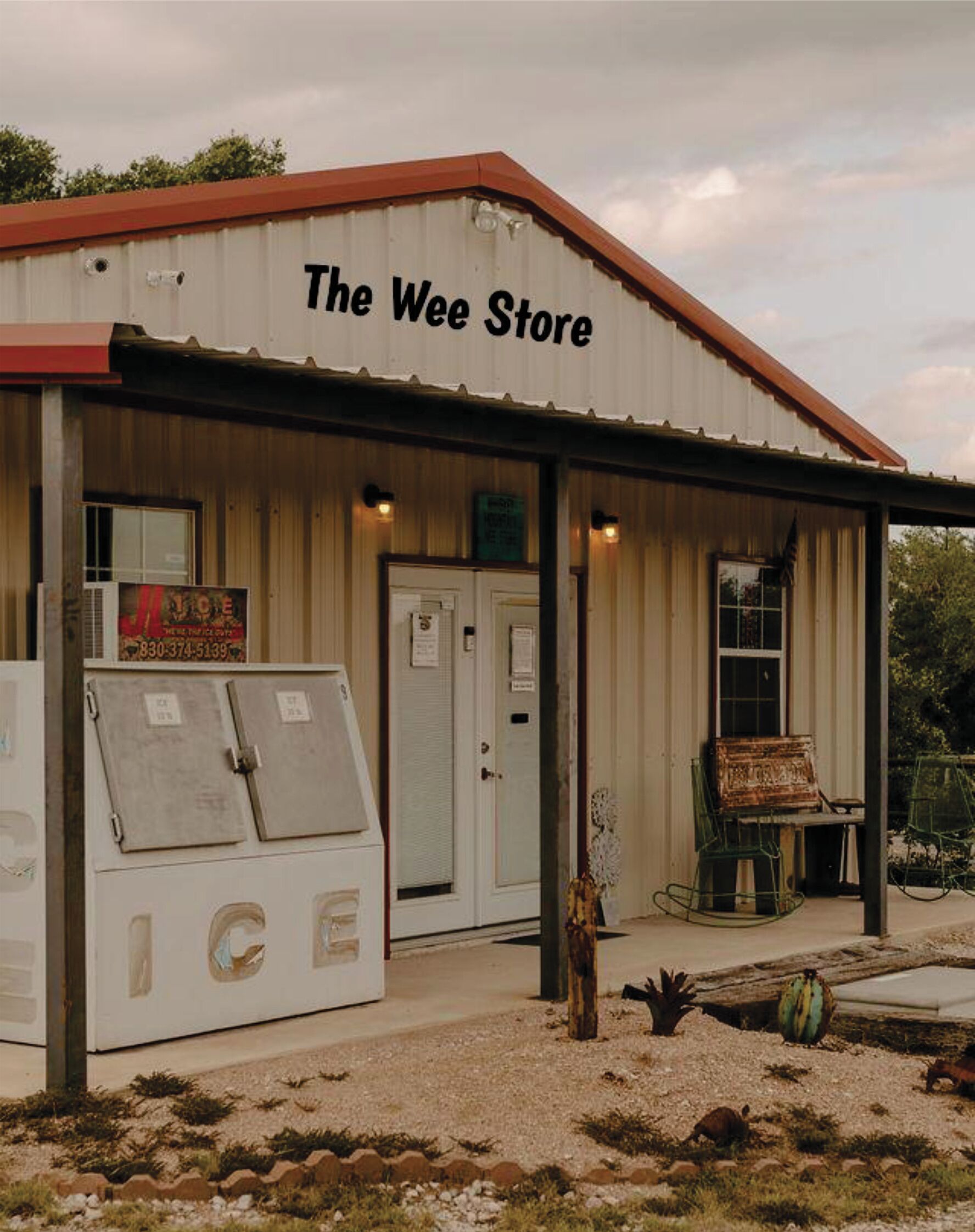 Un bâtiment portant l’inscription « The Wee Store », avec un toit en métal, une porte et un petit espace porche aménagé avec des chaises.