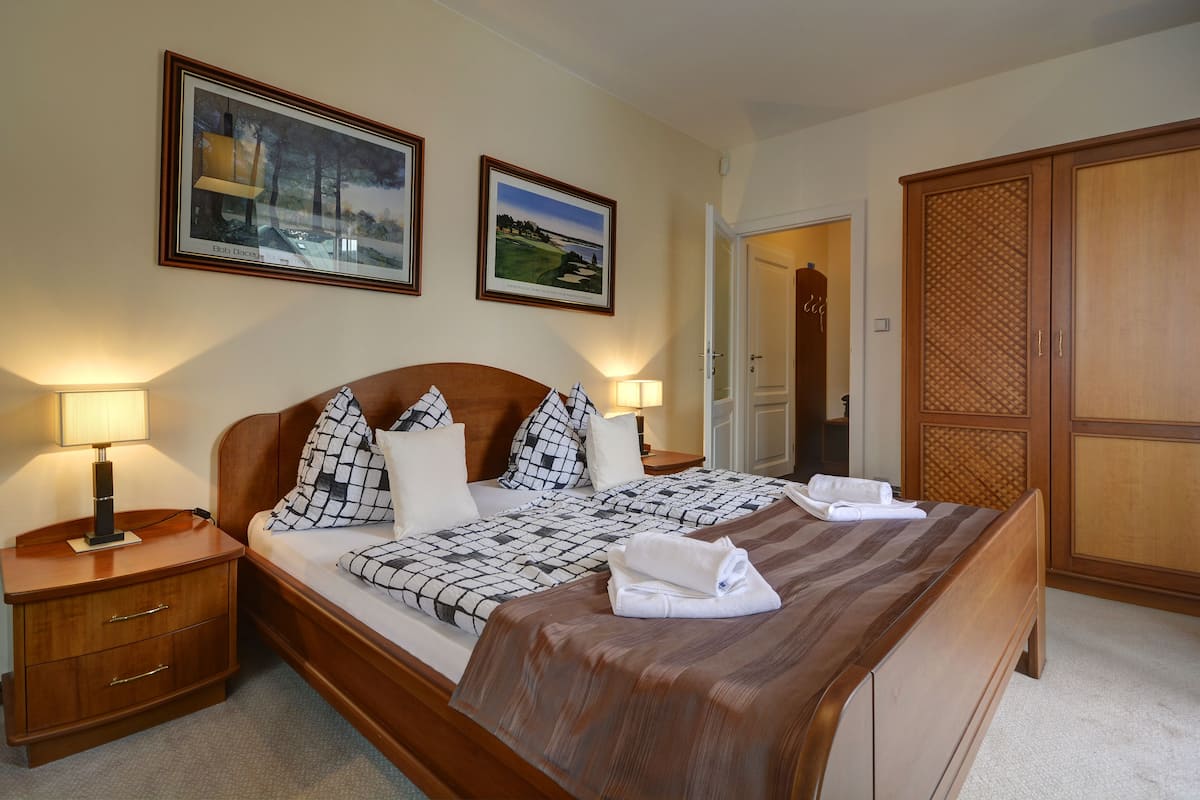 A neatly made bed with a checkered bedspread, two pillows, and a folded towel. There are two bedside lamps, a wooden nightstand, and a framed picture on the wall.