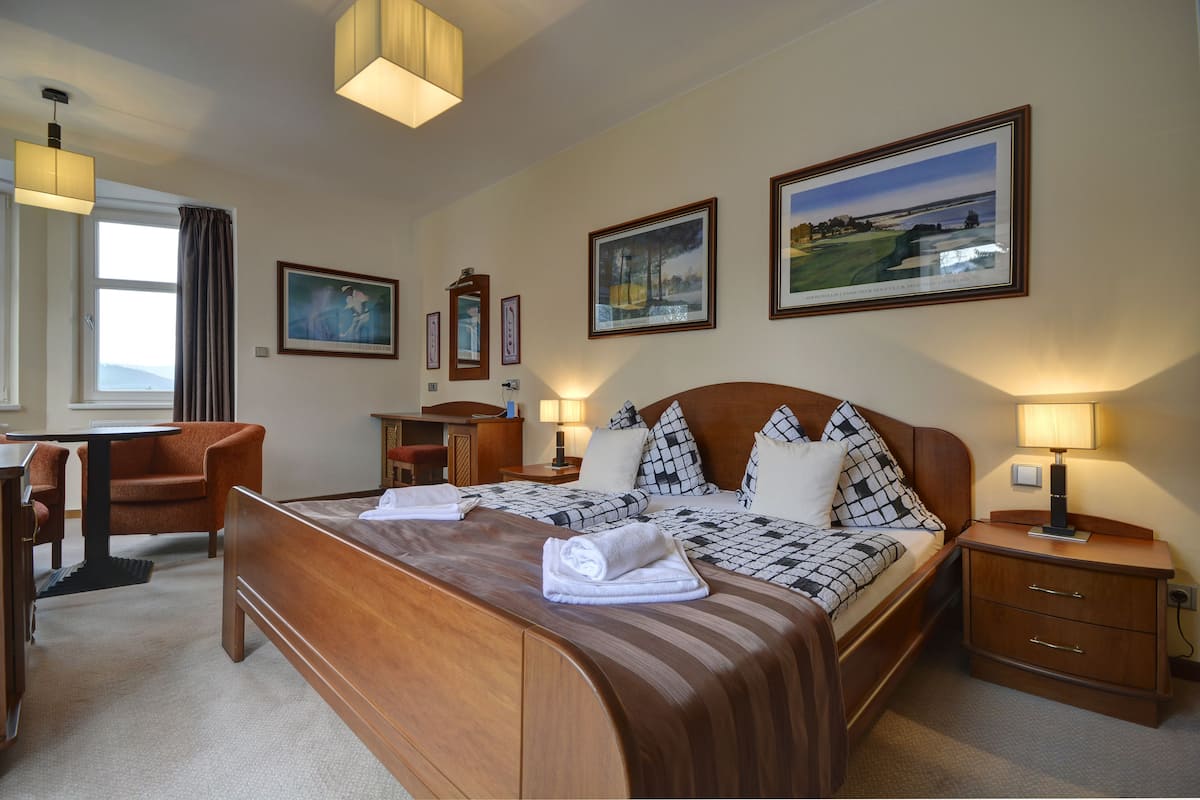 A hotel room with a large bed, a desk, and a chair. There are framed pictures on the wall and a window with curtains.