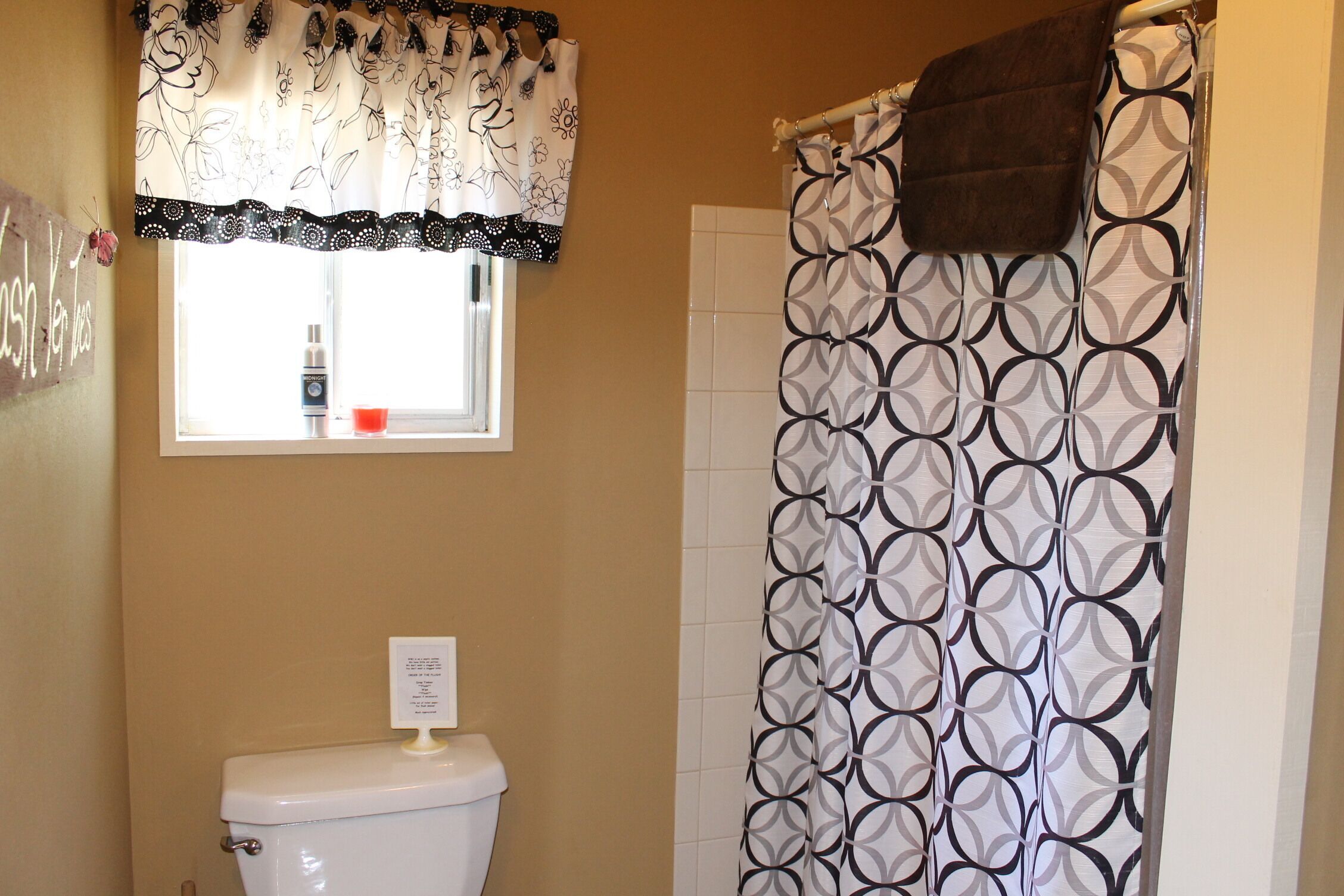 Une salle de bain comprenant des toilettes, une fenêtre agrémentée d’un rideau décoratif et un rideau de douche orné d’un motif géométrique.