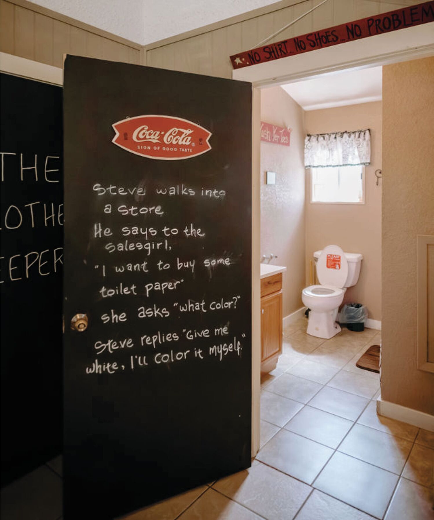A bathroom with a toilet, a sink, and a window with a curtain. There is a sign above the door that says "NO SHIRT NO SHOES NO PROBLEM".
