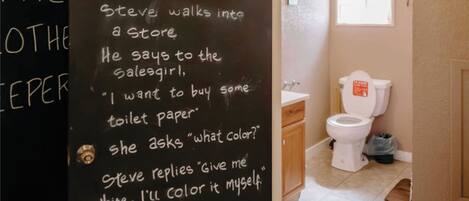 A bathroom with a toilet, a sink, and a window with a curtain. There is a sign above the door that says "NO SHIRT NO SHOES NO PROBLEM".
