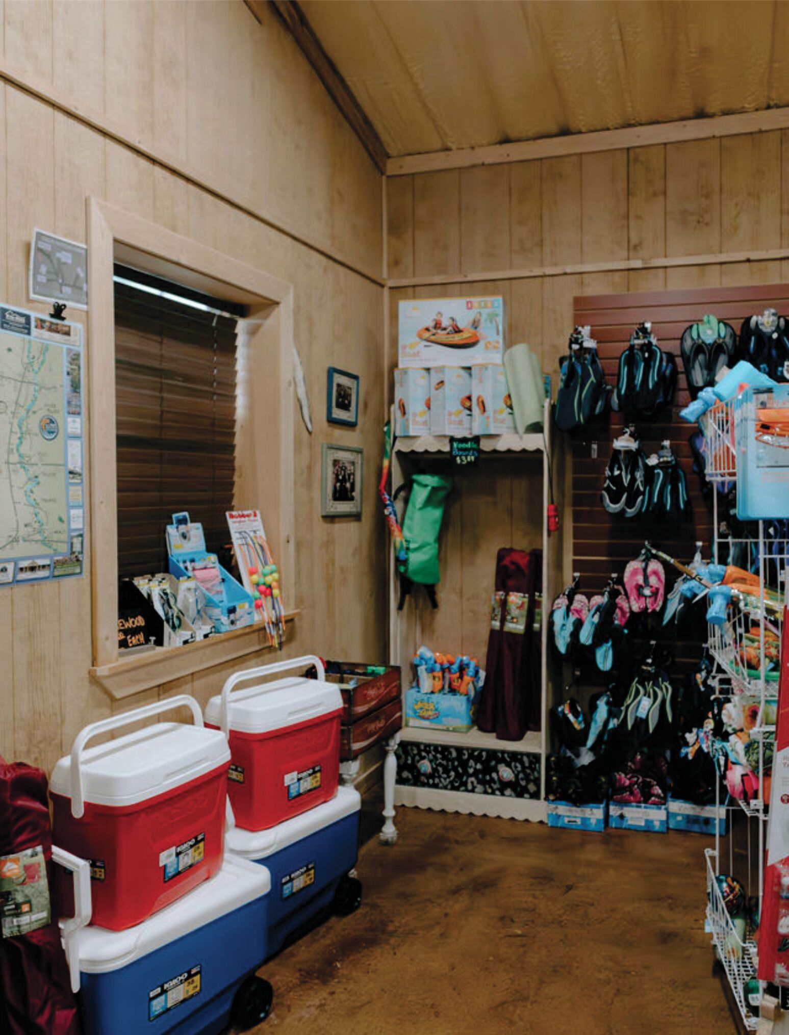 A room with wooden walls, a window, and shelves stocked with various items including coolers, bags, and children's toys.