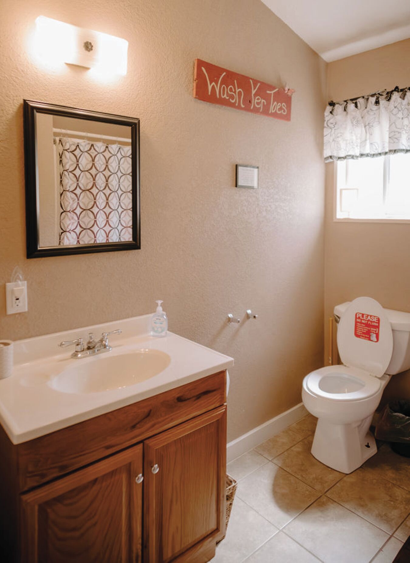 A bathroom with a wooden vanity, a sink, a mirror, a toilet, and a window with a curtain.