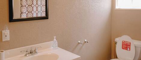 A bathroom with a wooden vanity, a sink, a mirror, a toilet, and a window with a curtain.