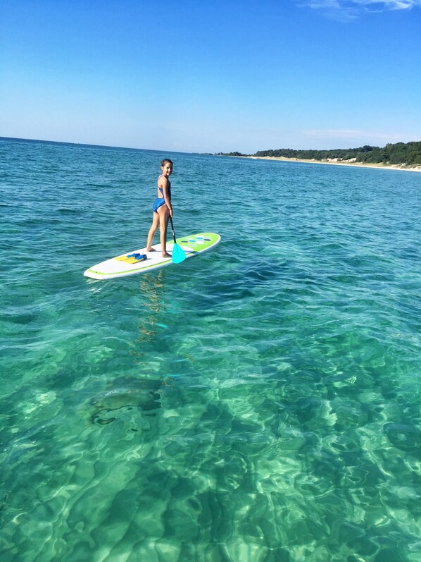 Una persona haciendo paddleboard en aguas claras y poco profundas, con una playa y árboles al fondo.