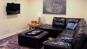 A living room with a black leather sofa, a flat-screen TV mounted on the wall, a side table with a lamp, and a framed cityscape picture. - Dartt's Park Garden View - 1 Bedroom (Owatonna)