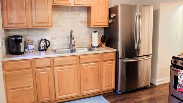 A kitchen with wooden cabinets, a stainless steel refrigerator, a coffee maker, and a sink. - Dartt's Park Garden View - 1 Bedroom (Owatonna)
