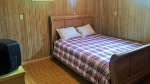 A single-bed room with wooden panel walls, a plaid bedspread, and a small window.