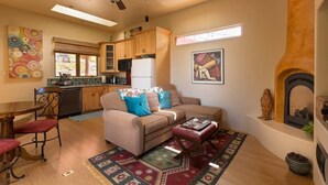 A cozy living room with a sofa, coffee table, and fireplace. There is a kitchen in the background with wooden cabinets and a refrigerator.