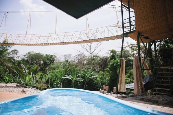 A swimming pool with a rope bridge overhead, surrounded by lush greenery and a thatched-roof structure.