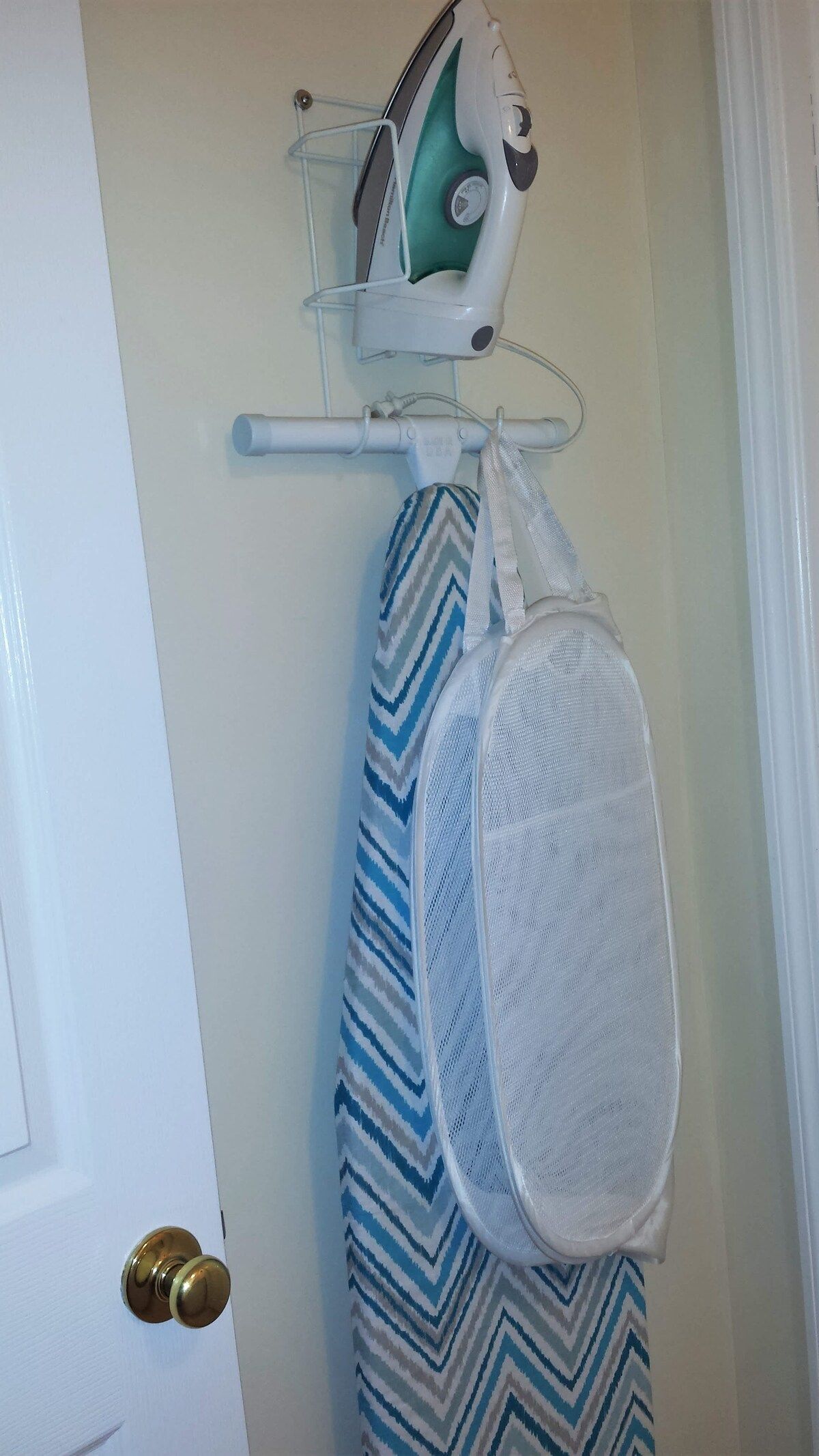 Een deur, een witte muur, een hangende strijkplank met een strijkijzer en een witte netmand voor wasgoed.