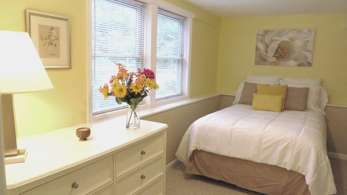 A bedroom with a bed, a dresser, a window with blinds, and a vase of flowers.