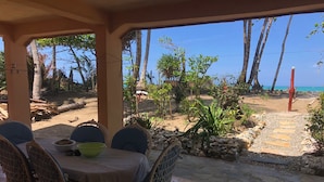 Een terras met rieten stoelen en een tafel, met uitzicht op een strand met palmbomen en een heldere lucht.