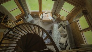 Interior de una cabaña de madera con una escalera en espiral, una mesa de comedor con sillas y un amplio ventanal con vistas al exterior.