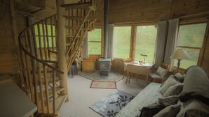 Interior de una cabaña con escalera espiral, estufa de leña, zona de comedor y grandes ventanales.