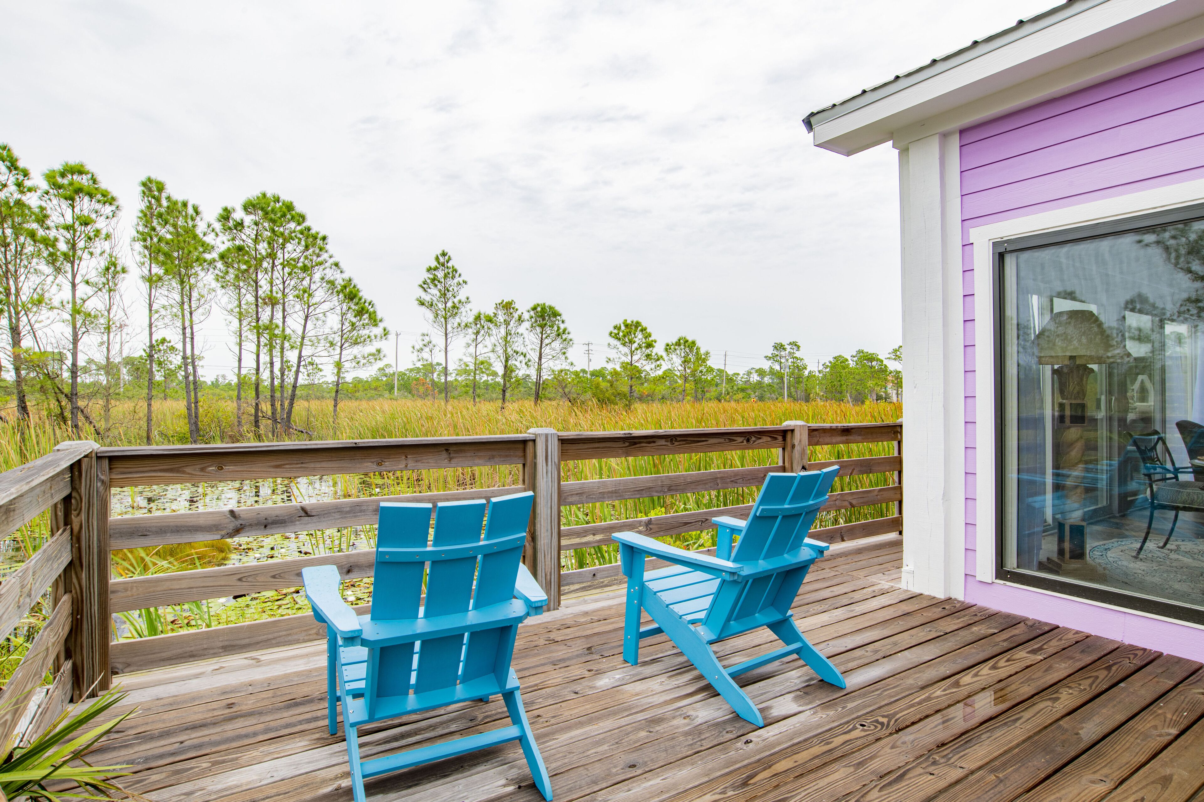 A wooden deck with two blue Adirondack chairs overlooking a marshy area with trees and water.