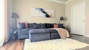 Un salon avec un canapé d’angle gris, un tapis blanc à poils longs et des œuvres d’art abstraites accrochées au mur.