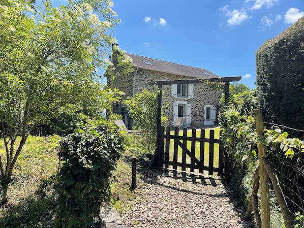 A stone house with a wooden gate, surrounded by greenery and a clear sky.