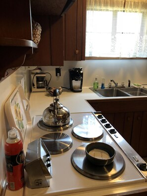 A kitchen with a stove, a coffee maker, a sink, and a window with lace curtains. - Historic Victoria Lea Guest Cottage In Downtown Tryon, NC (Tryon)