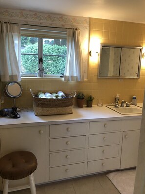 Een badkamer met een grote spiegel, een wastafel, een raam met gordijnen, een mandje met toiletartikelen en een krukje.