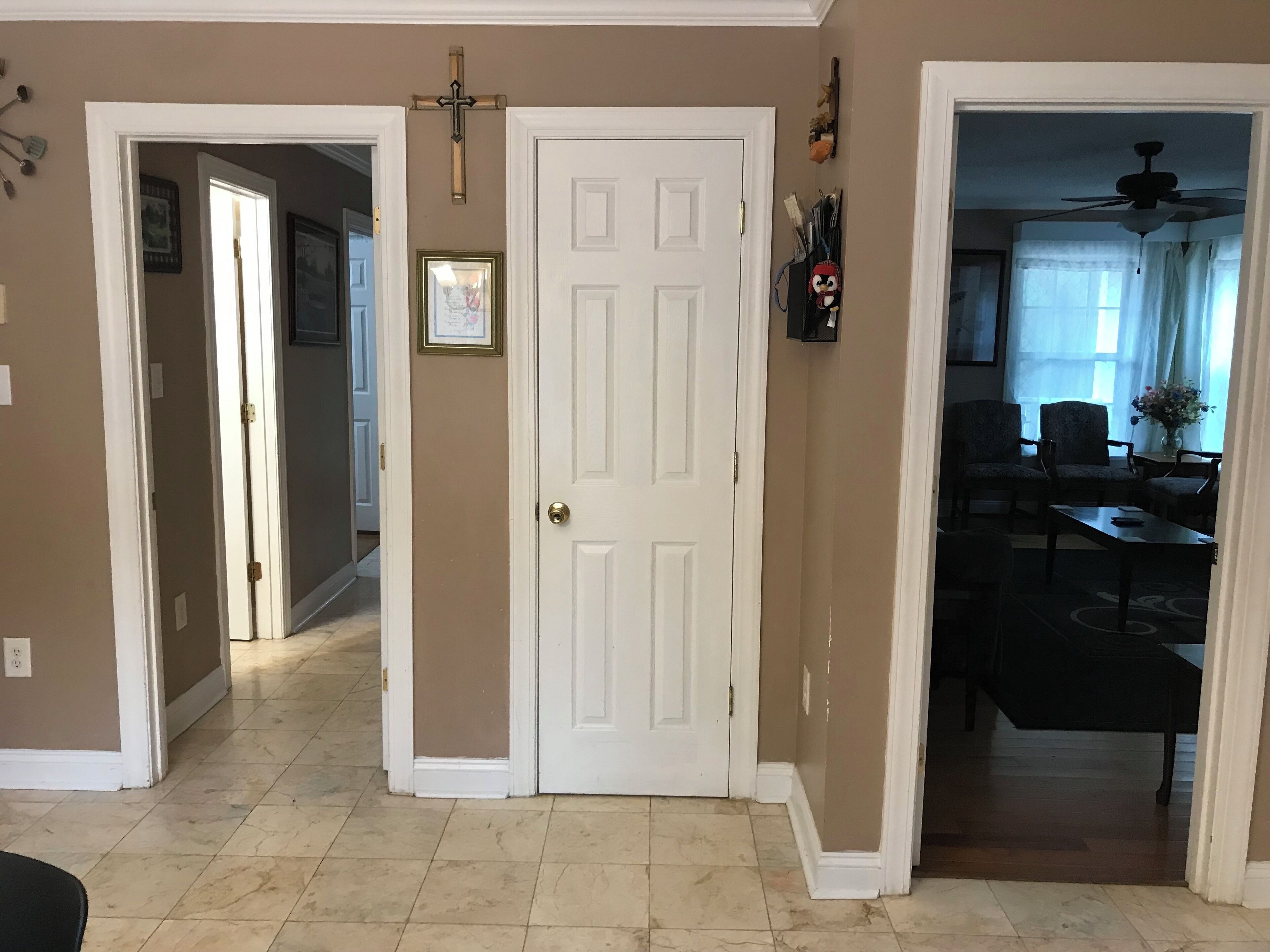 A hallway with a white door, beige walls, and a tiled floor. There are two chairs and a table in the adjacent room.